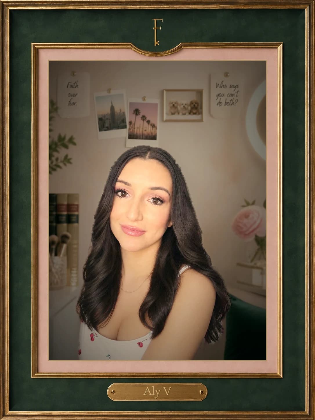 Aly V, smiling, her long dark hair over her shoulders, in a white top with cherries — her own photograph, set in a blush-pink studio: pinned to the wall behind her, a picture of the New York skyline, palm trees at a Los Angeles sunset, a framed photo of three small dogs, and notes reading “Faith over fear” and “Who says you can’t do both?”; law books and makeup brushes to one side, a pink peony and a ring light to the other — in a carved green-and-gold frame with a brass nameplate carrying her name.