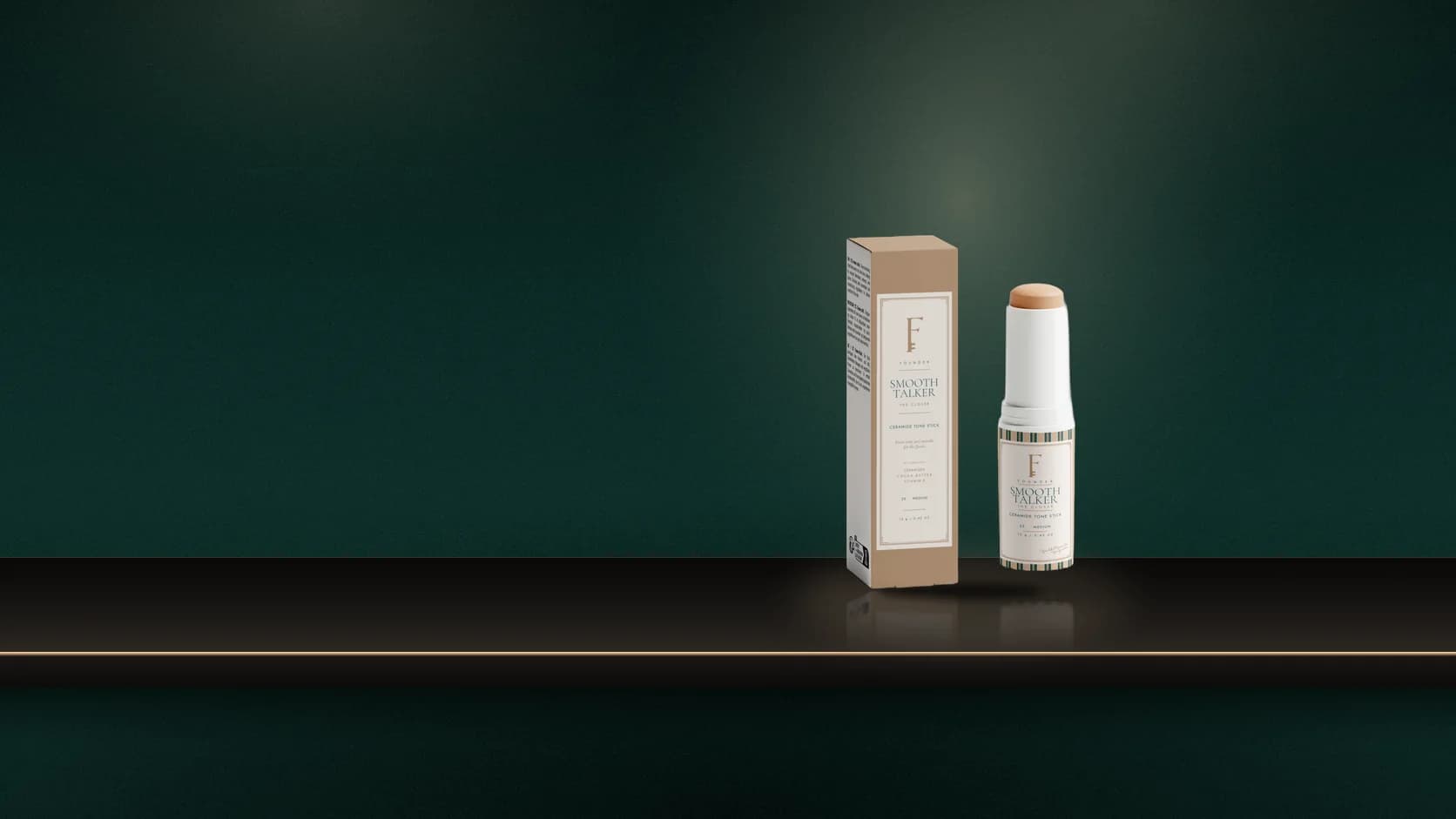 The Smooth Talker tone stick and its carton on a brass side table in the FOUNDER dressing room, a swatch of 25 MEDIUM on a stone dish, a woman blending it at her cheek in the mirror.